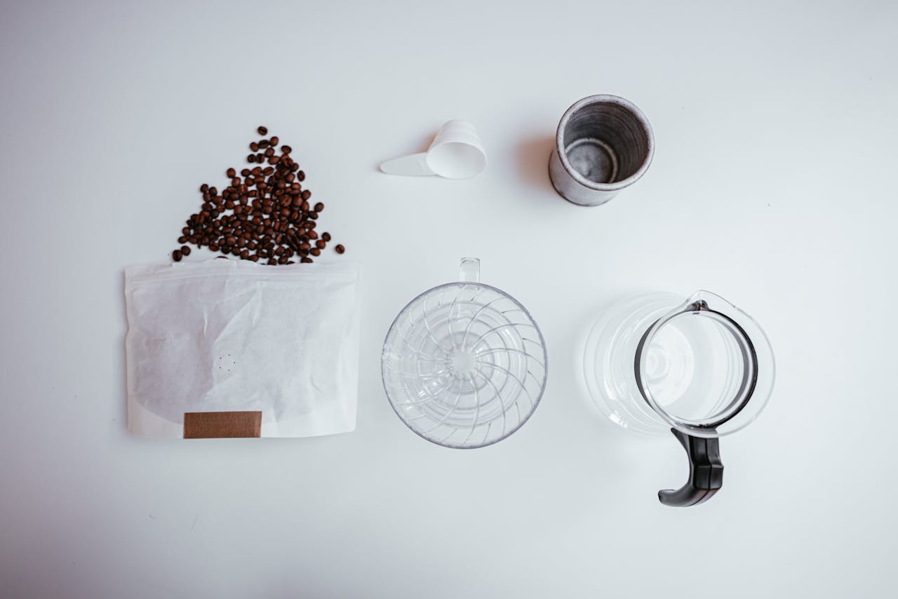 Top view of a minimalist coffee brewing setup with beans, dripper, and coffee pot on a white background.